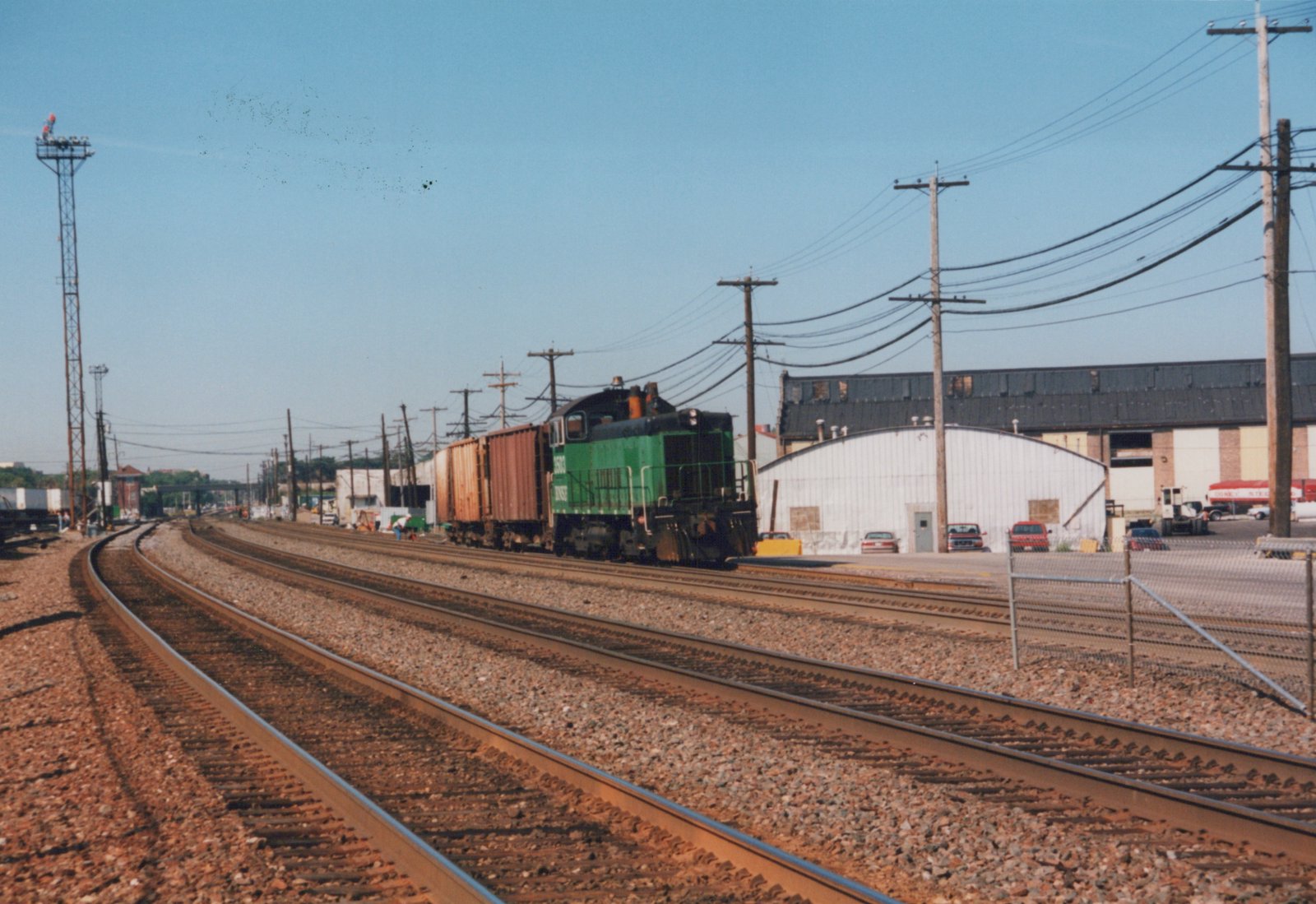 A green and black diesel locomotive with the letters BNSF on its side is moving along a set of railroad tracks, pulling several boxcars. The background shows industrial buildings and a clear blue sky. Several cars are parked nearby.