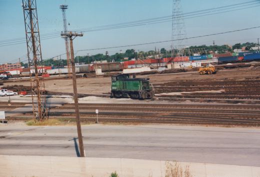 A green BNSF diesel locomotive numbered 3631 sits idle in a large rail yard with multiple tracks, freight containers, and a yellow construction vehicle in the background.
