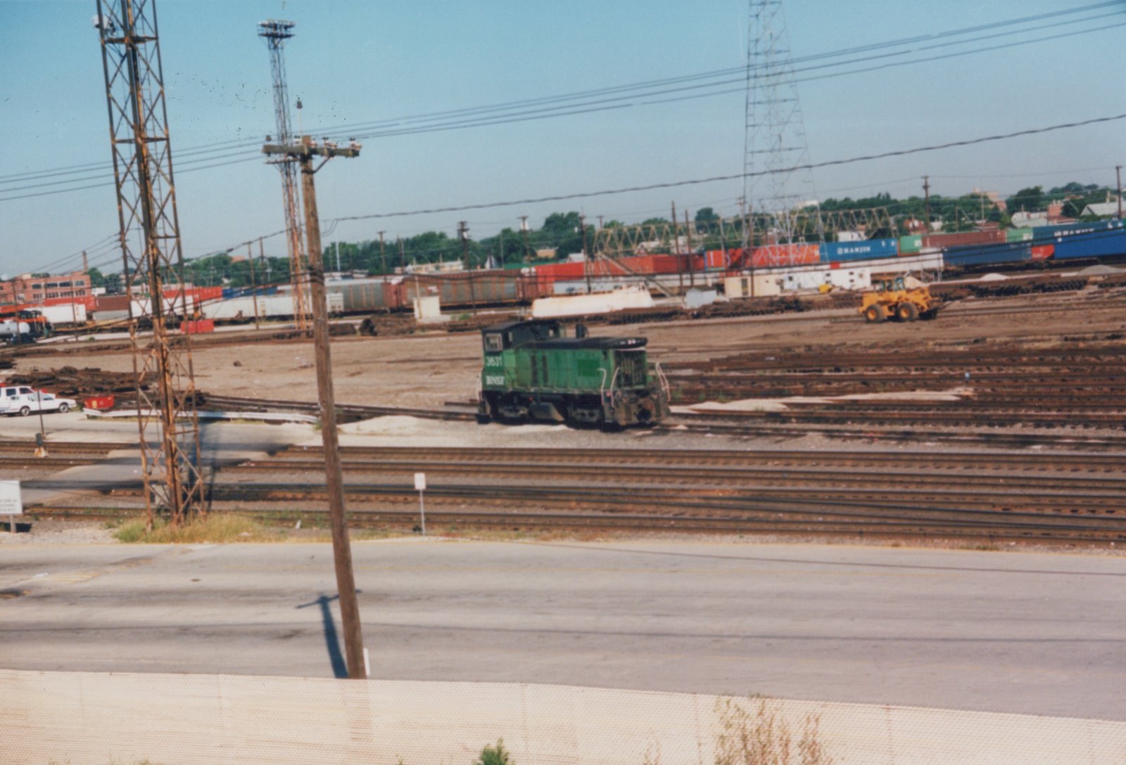 A green BNSF diesel locomotive numbered 3631 sits idle in a large rail yard with multiple tracks, freight containers, and a yellow construction vehicle in the background.