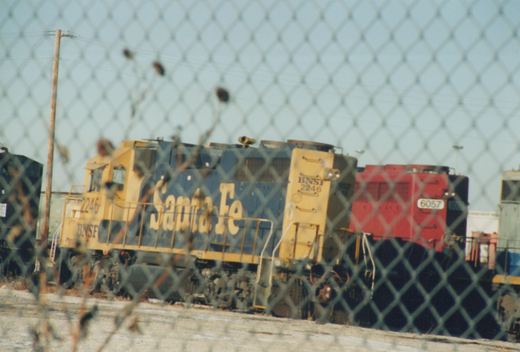 Two diesel locomotives, one blue and yellow with BNSF 2246 and Santa Fe lettering, the other red and black with BNSF 6057, are parked in a yard behind a chain-link fence on a clear winter day.