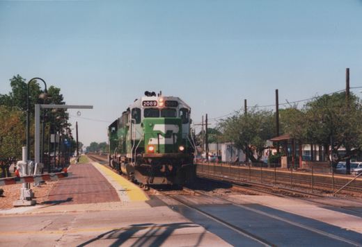 A green and white Metra locomotive numbered 2089 is approaching a station platform on a sunny day, with passengers waiting and cars parked nearby.