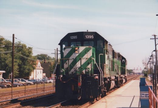 A green and white striped diesel locomotive numbered 1395 sits on the tracks beside a station platform with a 'Chicago Sun-Times' sign.