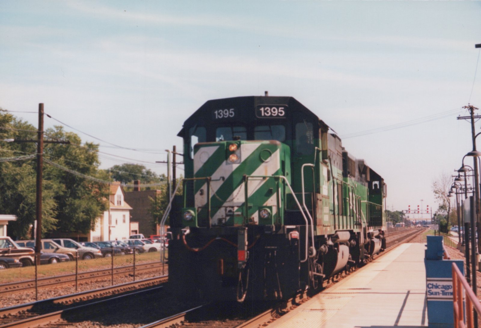 A green and white striped diesel locomotive numbered 1395 sits on the tracks beside a station platform with a 'Chicago Sun-Times' sign.