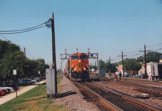 An orange BNSF locomotive numbered 1081 travels along tracks beside a roadside with parked cars, utility poles, and a few people nearby under a clear blue sky.