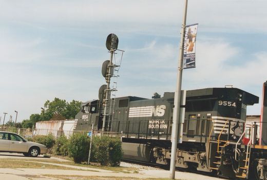 A black Norfolk Southern diesel locomotive numbered 9554 is parked beside a chain-link fence and green bushes, with a street pole bearing a Franklin Park banner in the foreground.