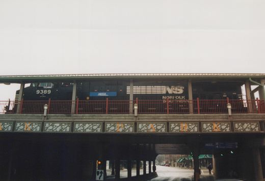 A black Norfolk Southern locomotive with the number 9389 and a Metra stripe is stopped on an elevated structure above a street; the structure has 'PARK' spelled out in letters below the tracks.
