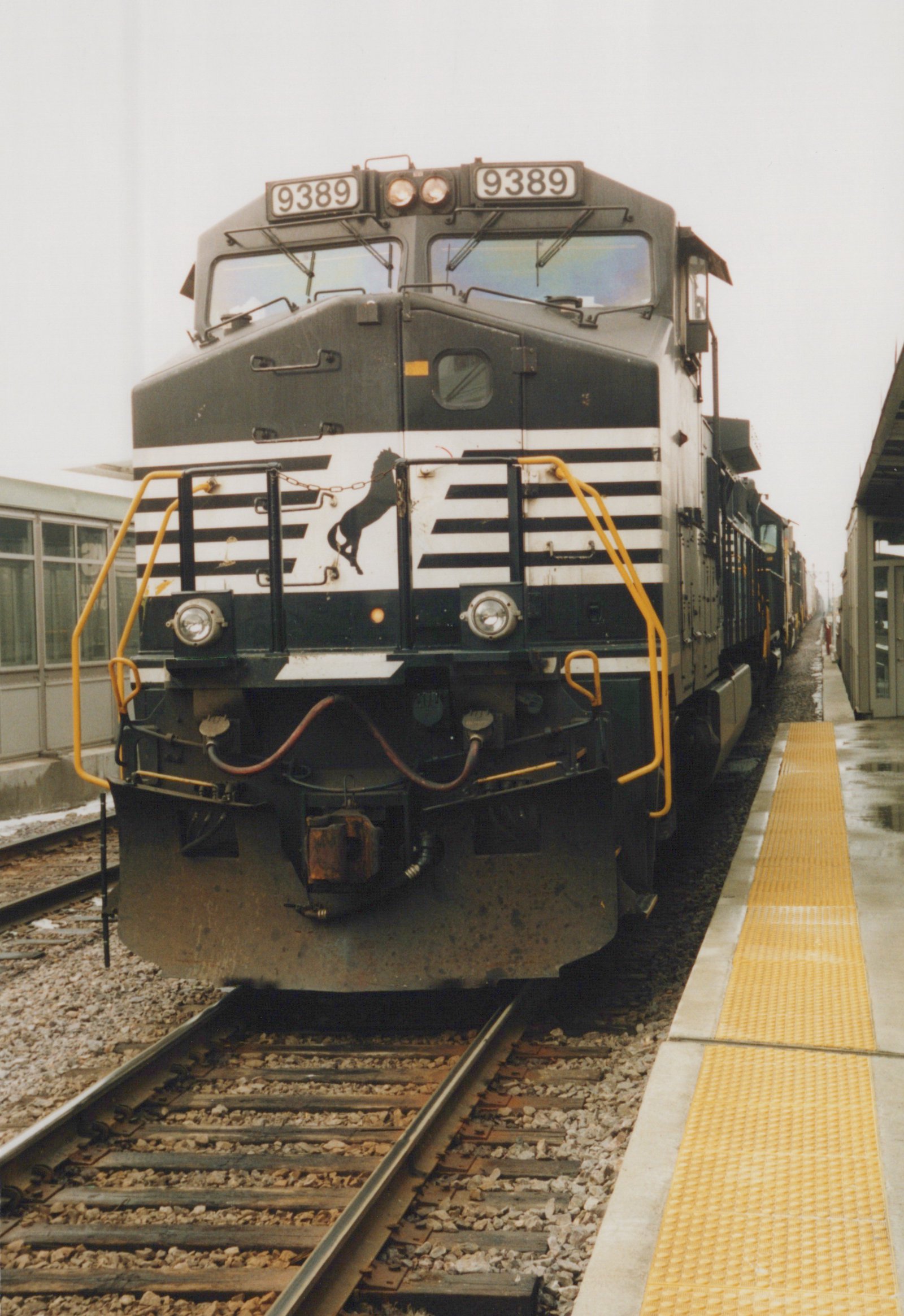 Front view of a black and white diesel locomotive with the number 9389 on its front, parked at a station platform with a yellow safety strip, on a cloudy winter day.