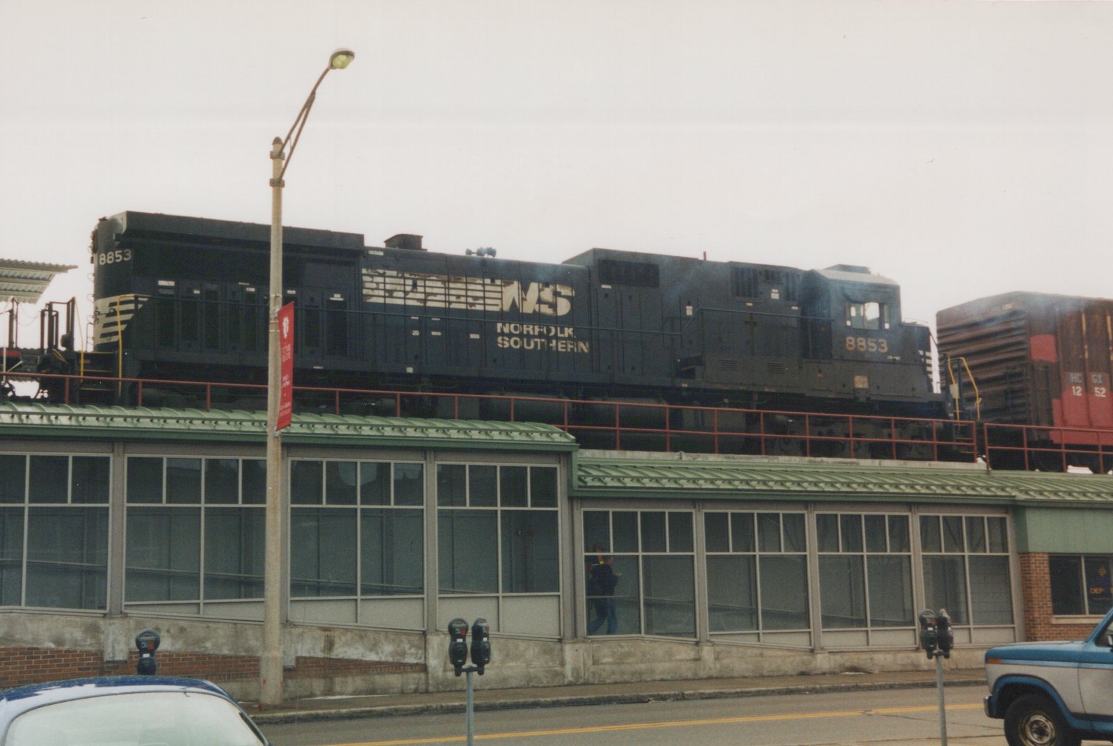A black Norfolk Southern diesel locomotive number 8853 on an elevated track above a street with cars and a building below, featuring a person walking on the sidewalk.