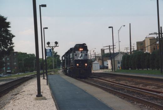 A black Chicago and North Western diesel locomotive numbered 5077 moves along tracks beside a station platform under an overcast sky.