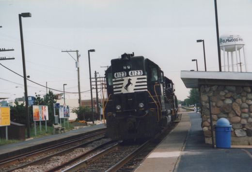A black diesel locomotive with white stripes and the number 4123 on the front is parked at a station platform next to a stone shelter, with a water tower labeled 'Elmwood Park' in the background.
