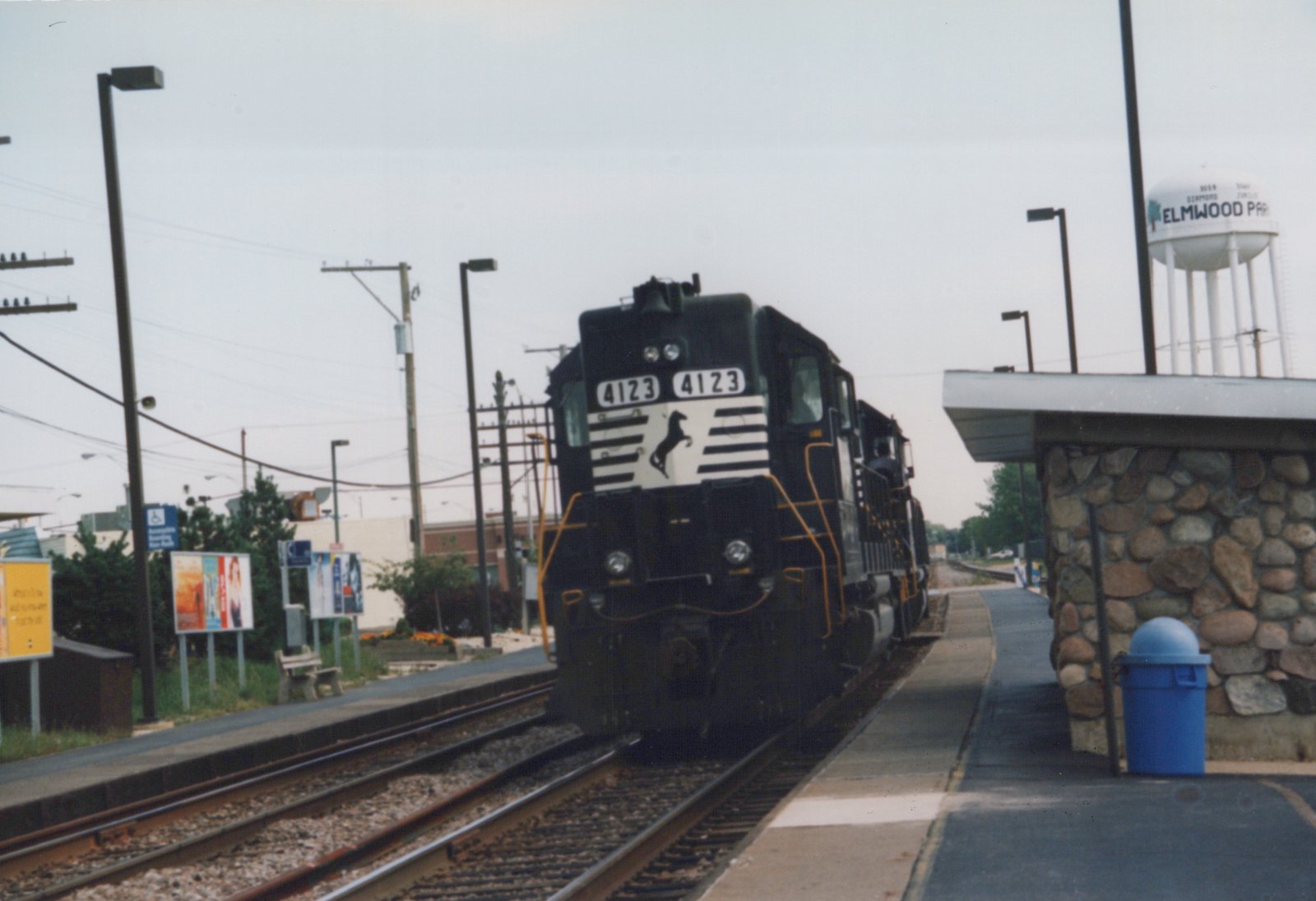 A black diesel locomotive with white stripes and the number 4123 on the front is parked at a station platform next to a stone shelter, with a water tower labeled 'Elmwood Park' in the background.