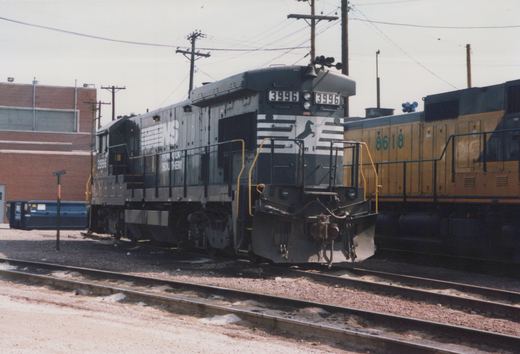 A black and white Norfolk Southern diesel locomotive numbered 3996 is shown from the front three-quarter view, parked on tracks in a yard with a yellow locomotive numbered 8618 visible behind it, under an overcast sky with some snow on the ground.