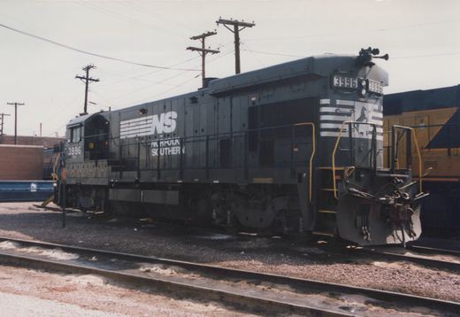 A gray Norfolk Southern diesel locomotive, number 3996, with white lettering and stripes, is parked on railroad tracks in a railyard with muddy ground and utility poles in the background.