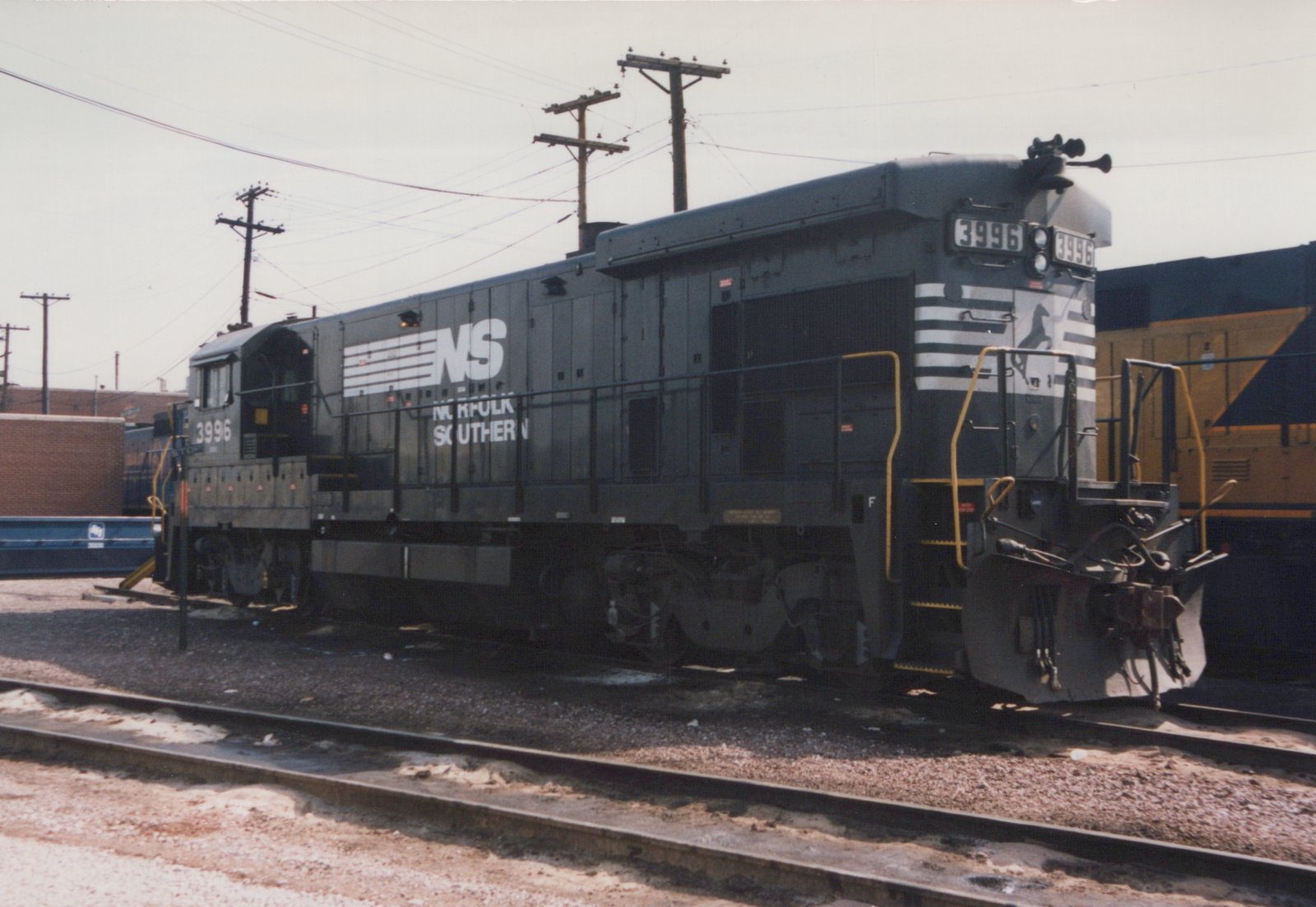 A gray Norfolk Southern diesel locomotive, number 3996, with white lettering and stripes, is parked on railroad tracks in a railyard with muddy ground and utility poles in the background.