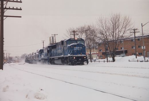 A blue Conrail GP38-2 diesel locomotive, number 5629, pulls a train of tanker cars along snow-covered tracks in a residential area, with bare trees and brick buildings in the background.