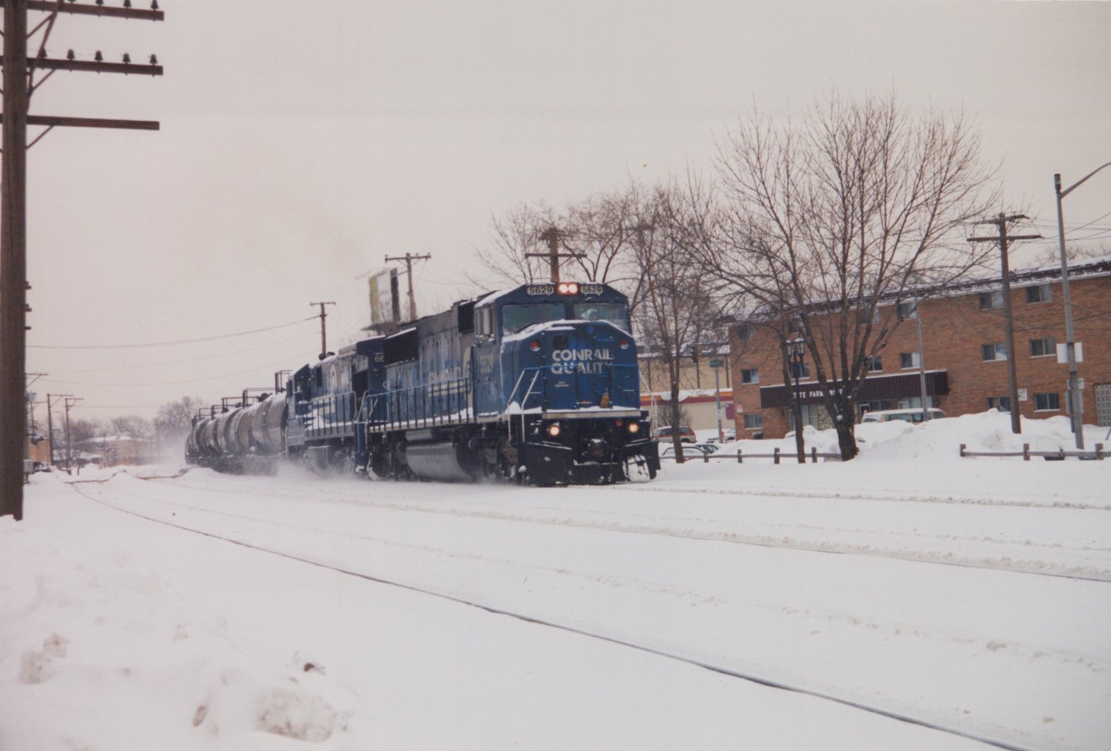 A blue Conrail GP38-2 diesel locomotive, number 5629, pulls a train of tanker cars along snow-covered tracks in a residential area, with bare trees and brick buildings in the background.