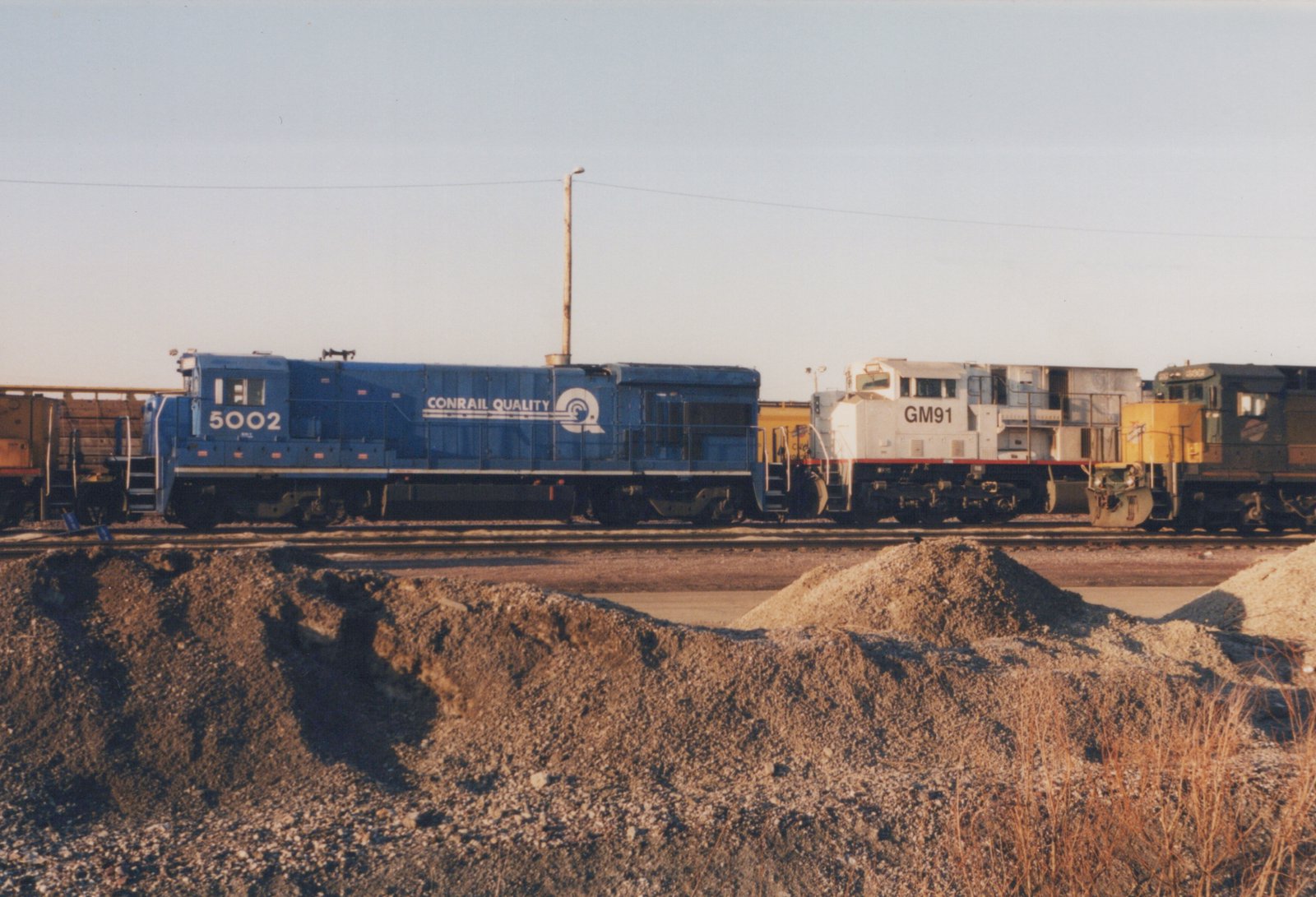 Two diesel locomotives, one blue with Conrail lettering and number 5002 and one white with GM91, are parked next to each other in a gravel rail yard under a clear sky.
