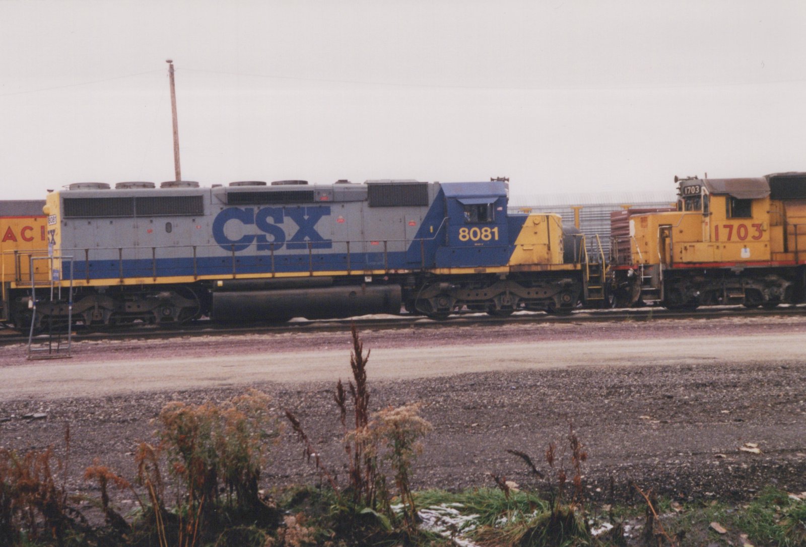 Side view of three diesel locomotives coupled together on tracks in a rail yard; the lead unit is blue and grey with 'CSX 8081' on the cab, the second is yellow with '1703', and the third is also yellow with 'ACI' on the side. Patches of snow are visible on the ground.