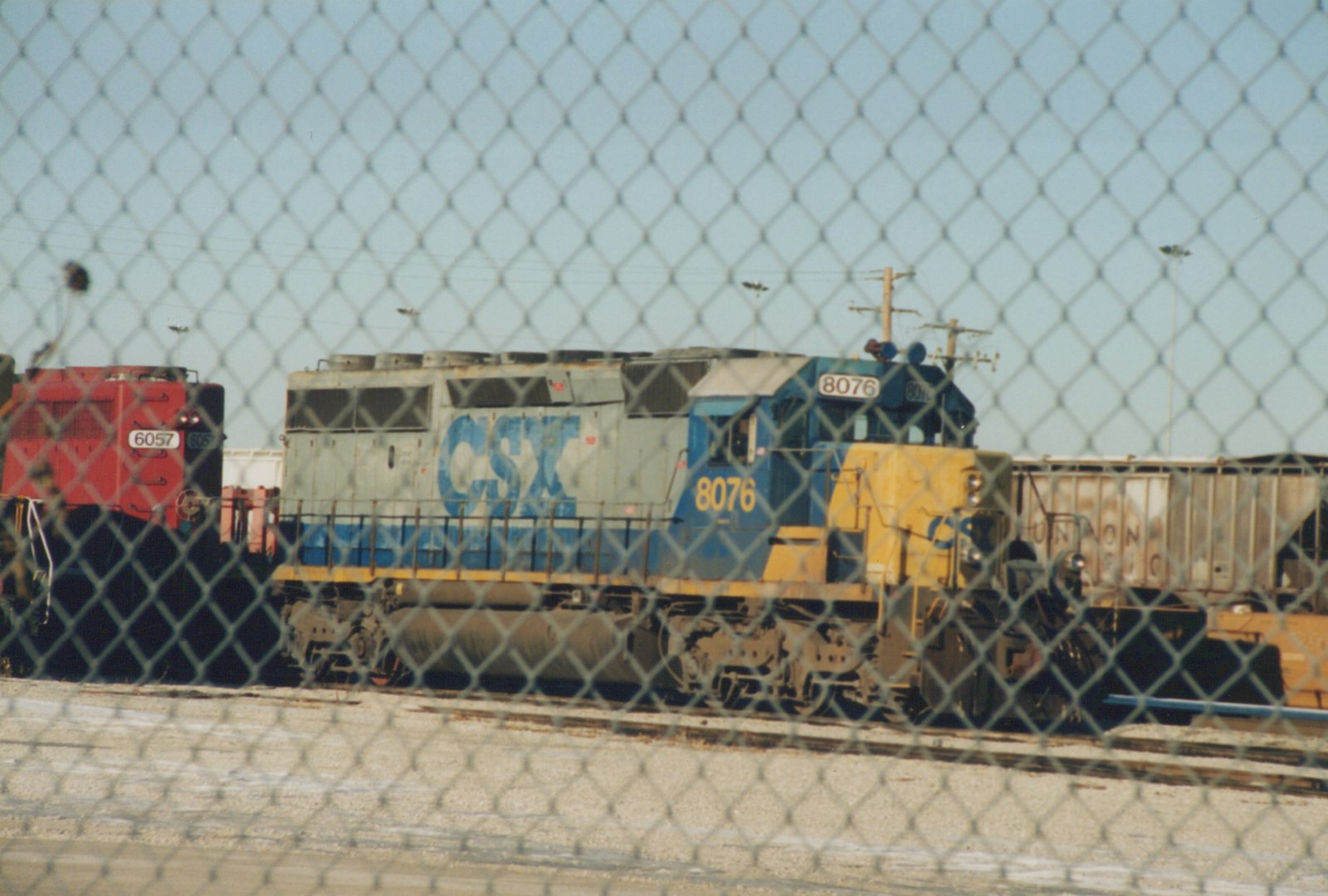 Two CSX diesel locomotives, numbers 8076 and 6057, are parked on tracks in a rail yard, seen through a chain-link fence under a clear sky.