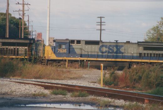 A blue, gray, and yellow CSX diesel locomotive, number 7535, stands on a set of railroad tracks beside overgrown vegetation and utility poles under an overcast sky.