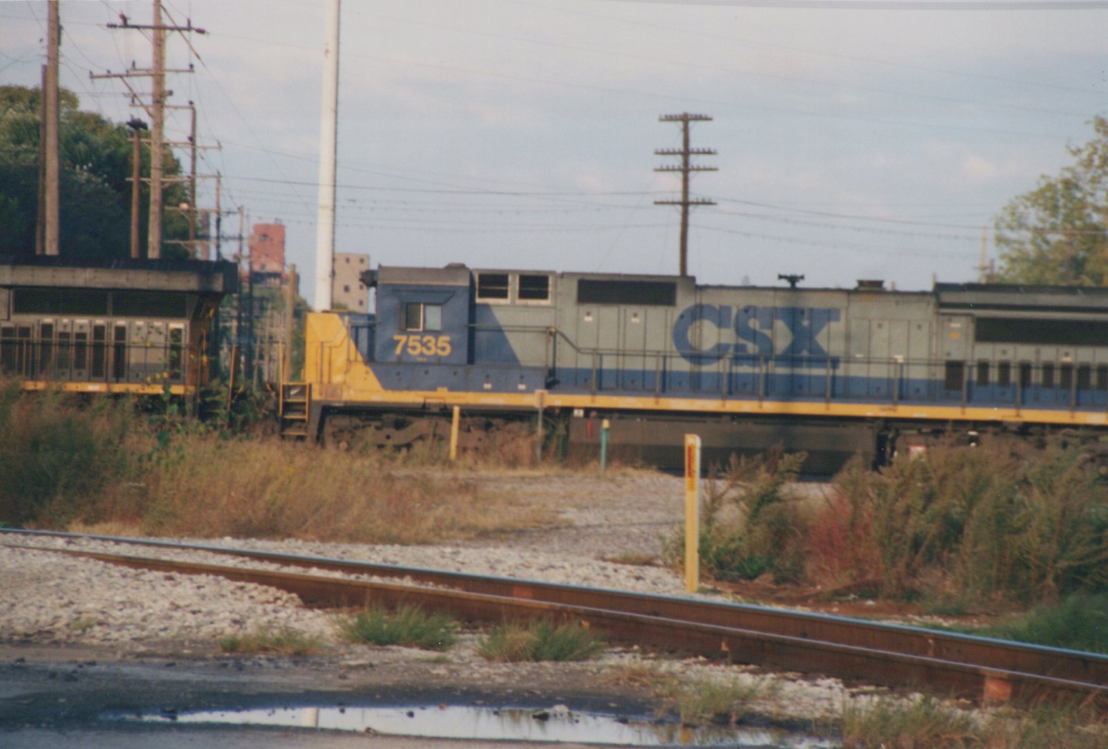 A blue, gray, and yellow CSX diesel locomotive, number 7535, stands on a set of railroad tracks beside overgrown vegetation and utility poles under an overcast sky.