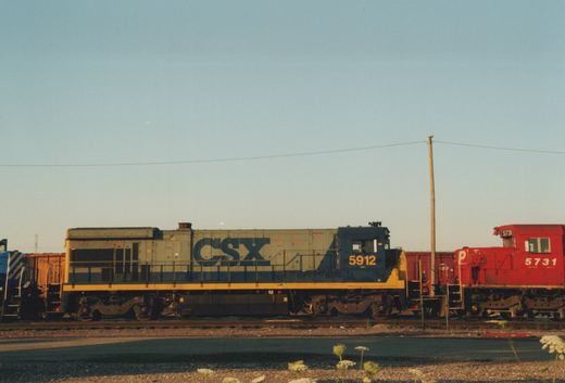 A side view of two diesel locomotives on parallel tracks: a gray and blue CSX locomotive numbered 5912 next to a red Pennsylvania Railroad locomotive numbered 5731, with a clear sky and a utility pole in the background.