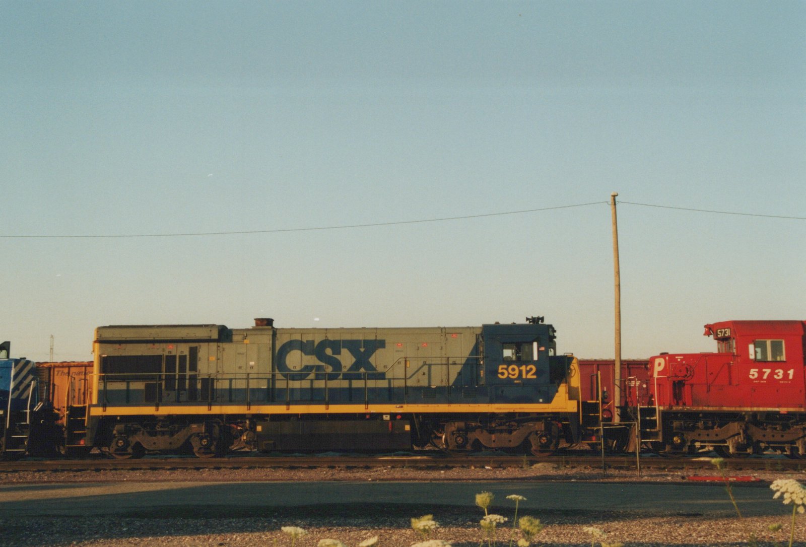 A side view of two diesel locomotives on parallel tracks: a gray and blue CSX locomotive numbered 5912 next to a red Pennsylvania Railroad locomotive numbered 5731, with a clear sky and a utility pole in the background.