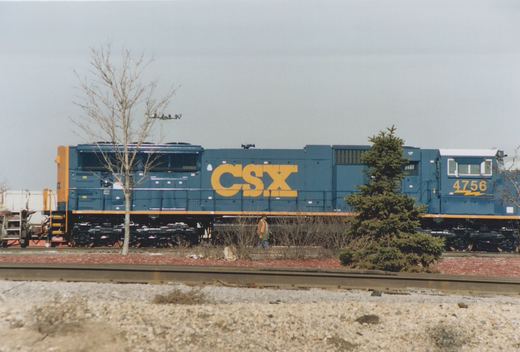 A side view of a blue and yellow CSX diesel locomotive with the number 4756, standing on tracks beside a bare tree and a person wearing a safety vest.