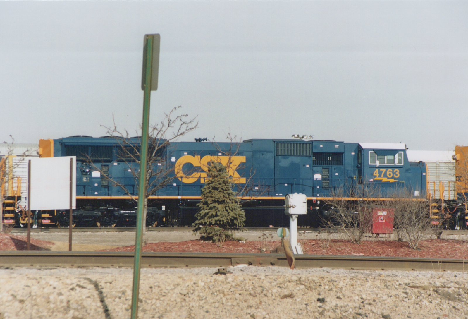 A blue and yellow CSX locomotive with the number 4763 on the cab side, seen from the side with bare trees and a cloudy sky in the background.