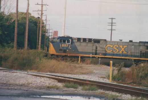 A blue, yellow and gray CSX Transportation diesel locomotive numbered 657 is seen on a railroad track next to overgrown vegetation and utility poles under an overcast sky.