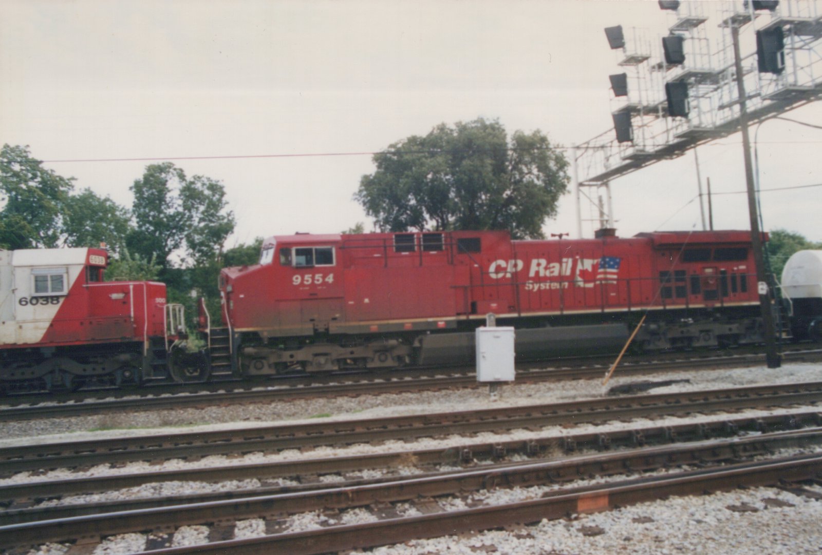 Two diesel locomotives, one red with CP Rail System lettering and number 9554, and one white and red with number 6038, are parked on parallel tracks in a yard under an overcast sky.