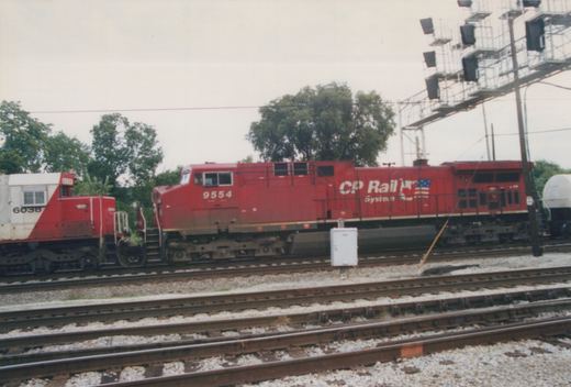 Two diesel locomotives, one red with CP Rail System lettering and number 9554, and one white and red with number 6038, are parked on parallel tracks in a yard under an overcast sky.