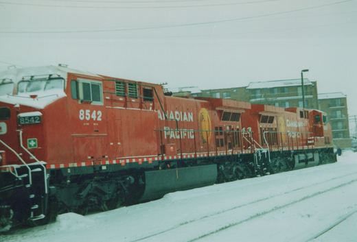 A bright red Canadian Pacific diesel locomotive, number 8542, stands on a snow-covered track with a second unit behind it, against a backdrop of a brick apartment building on a cloudy, snowy day.