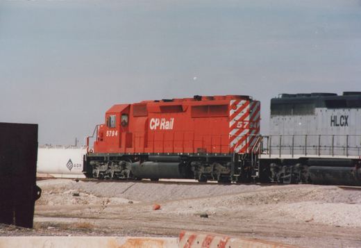 A side view of a bright red CP Rail diesel locomotive numbered 5794, leading a train with a gray HLCX locomotive behind, at a gravel grade crossing under an overcast sky.