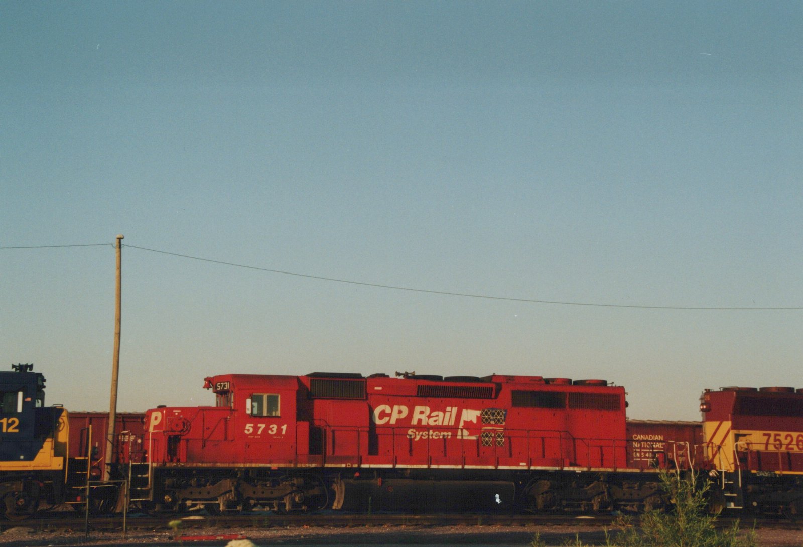 A red CP Rail System GP9 locomotive numbered 5731 is seen in a yard, with a blue and yellow locomotive and a Canadian National freight car in the background.