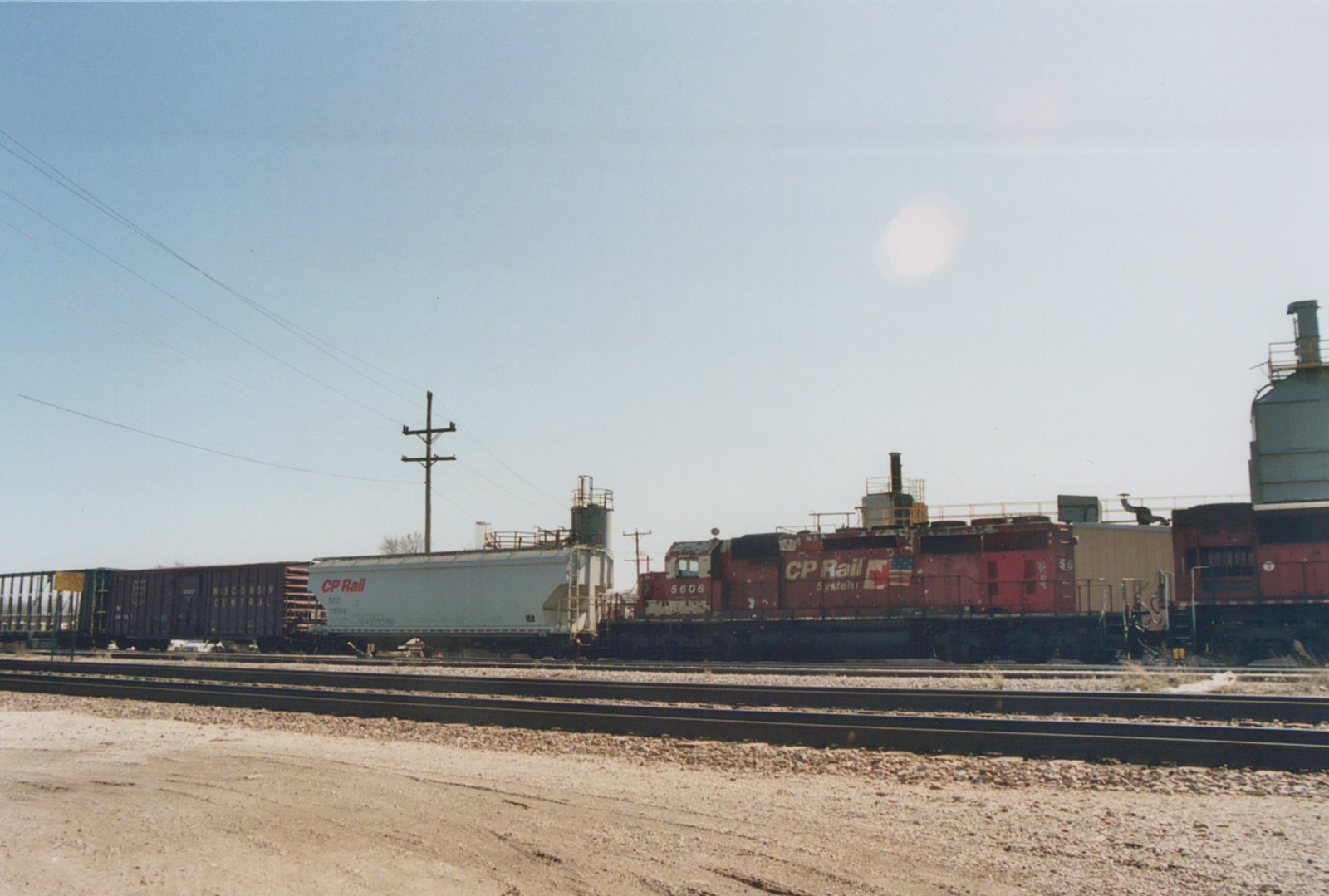 A red CP Rail System diesel locomotive numbered 5606 is shown on railroad tracks in a yard, pulling freight cars including a white covered hopper and a dark Wisconsin Central boxcar under a clear sky.