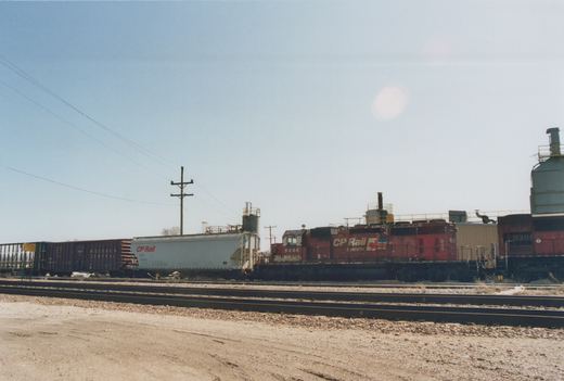 A red CP Rail System diesel locomotive numbered 5606 is shown on railroad tracks in a yard, pulling freight cars including a white covered hopper and a dark Wisconsin Central boxcar under a clear sky.