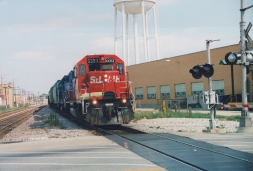 A red locomotive with 'StL&H' lettering and the number 5593 on the front travels on tracks through a grade crossing, with a water tower and brick building in the background.