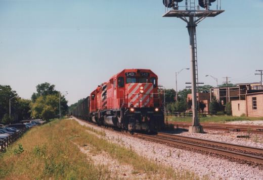 A red Canadian Pacific diesel locomotive, number 5401, with white diagonal stripes on the front, is pulling a long line of freight cars on a railroad track under a clear blue sky, with trees and parked cars on the left and a brick building on the right.
