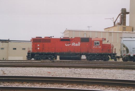 A red locomotive with 'CP Rail' on its side, numbered 3018, is on railroad tracks next to industrial buildings.