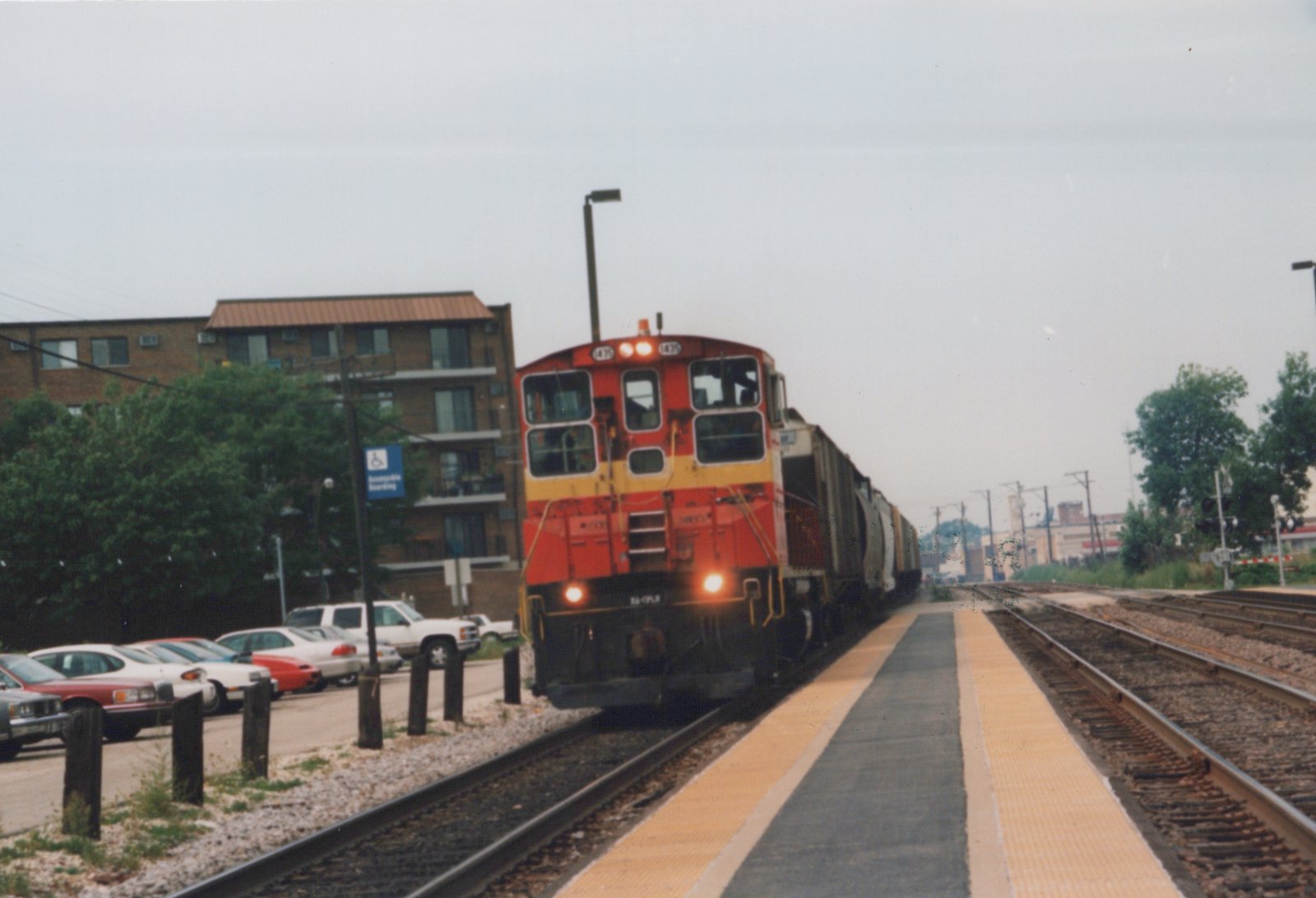 A red and yellow Canadian Pacific locomotive, number 475, is on the tracks at a station platform with parked cars and a brick apartment building in the background.