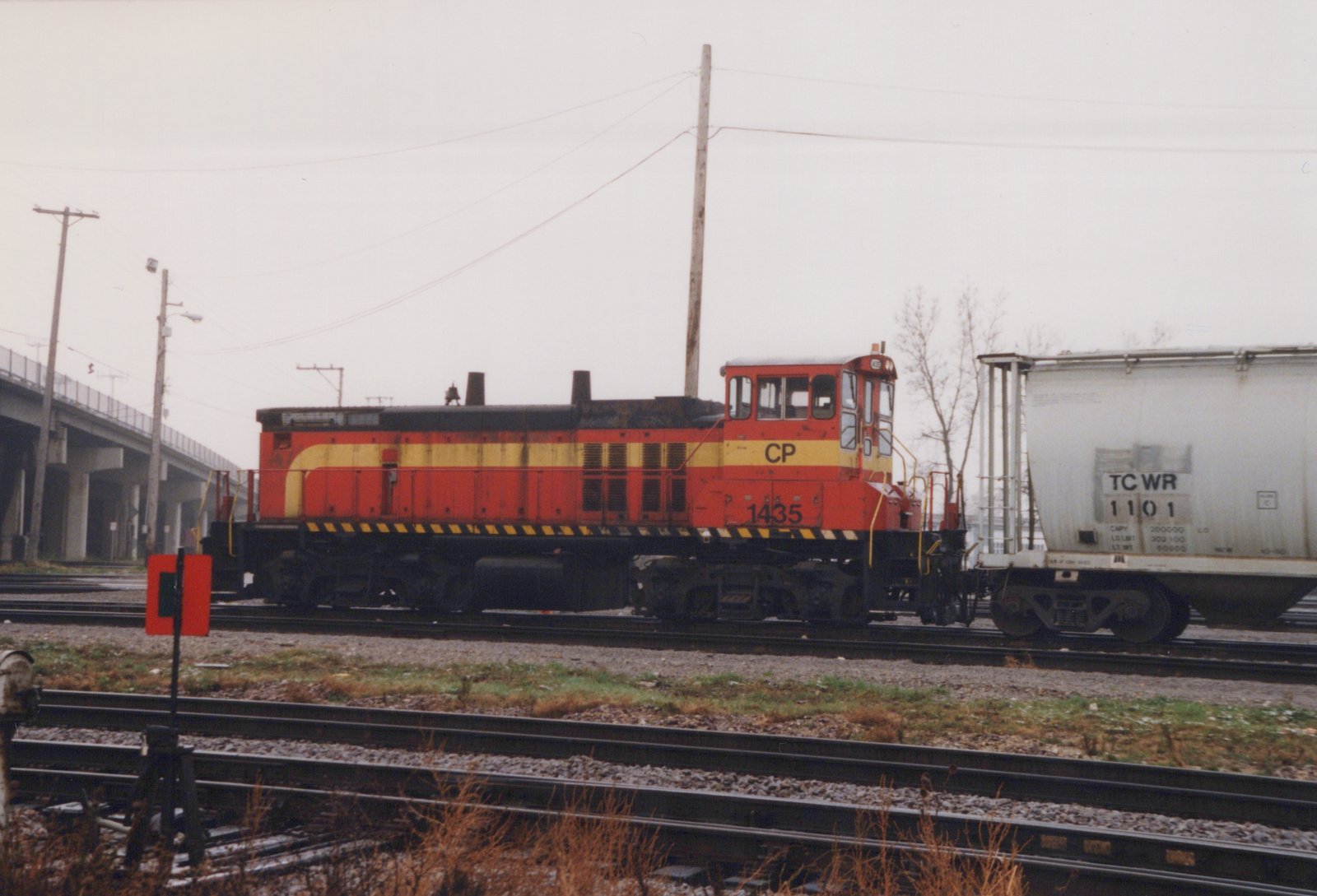 A red and yellow Canadian Pacific diesel locomotive numbered 1435 is parked on railroad tracks in a yard, next to a white freight car, with a bridge and utility poles visible in the background.