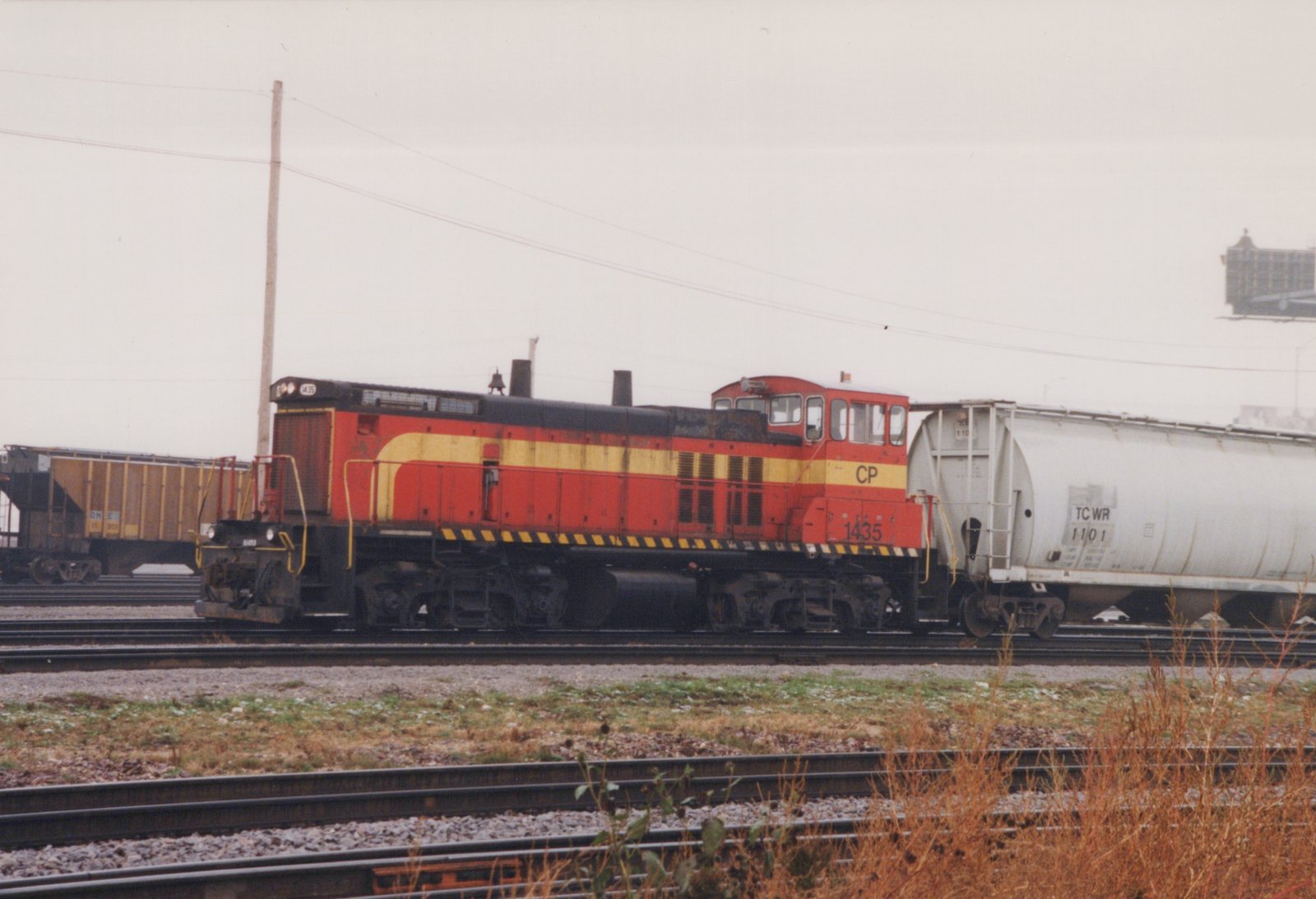 A red and yellow Canadian Pacific diesel locomotive, number 1435, is parked on tracks in a rail yard, coupled to a white tank car, with brown autumn weeds in the foreground under an overcast sky.