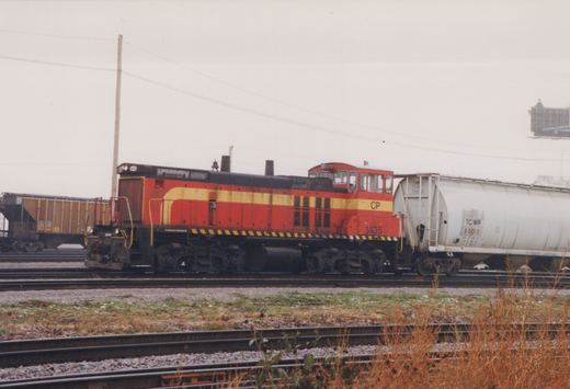 A red and yellow Canadian Pacific diesel locomotive, number 1435, is parked on tracks in a rail yard, coupled to a white tank car, with brown autumn weeds in the foreground under an overcast sky.