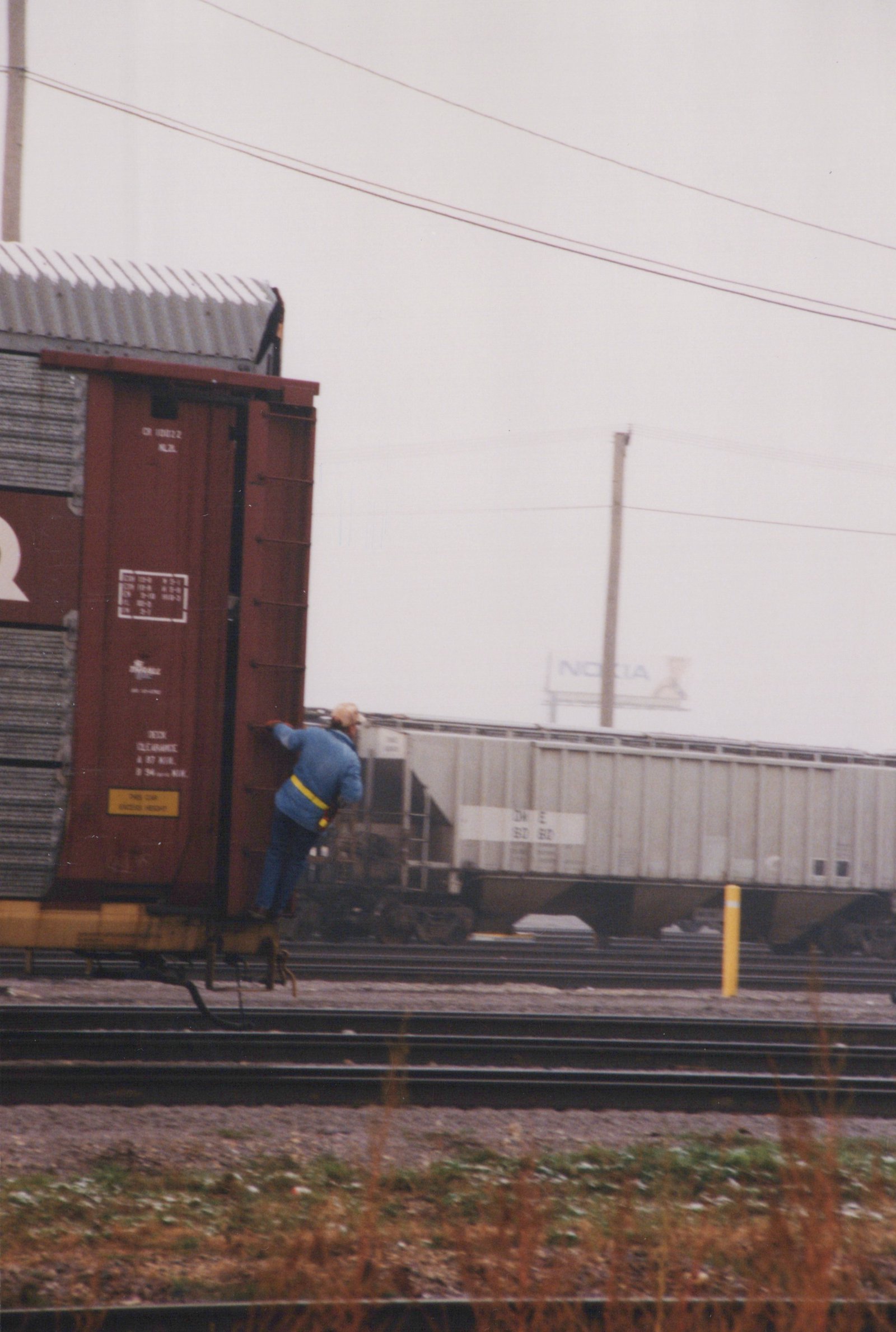 A person in blue clothing is climbing onto a red freight car in a rail yard with multiple tracks; a gray hopper car and a distant Nokia billboard are also visible.