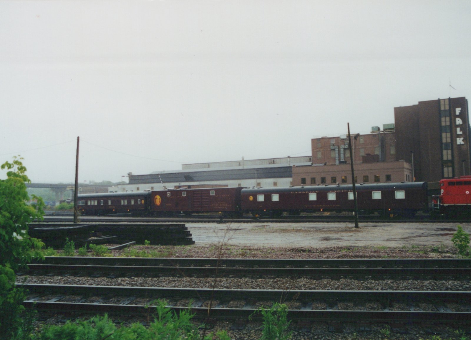 A side view of a Canadian Pacific train in a rail yard, featuring a red locomotive, a maroon sleeper car, and other passenger cars, with industrial buildings in the background under an overcast sky.