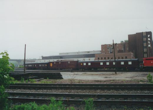 A side view of a Canadian Pacific train in a rail yard, featuring a red locomotive, a maroon sleeper car, and other passenger cars, with industrial buildings in the background under an overcast sky.