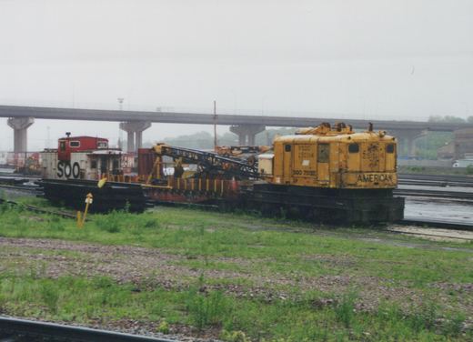An overcast day in a rail yard features a yellow American work unit, numbered 9500 250114, coupled to a red and white SOO caboose, with an elevated highway bridge in the background.