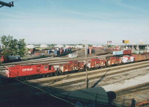 A red CP Rail locomotive numbered 1511 pulls a string of rusty and white SOO Line cabooses through a sprawling rail yard filled with freight cars and containers, with green trees and a clear blue sky in the background.