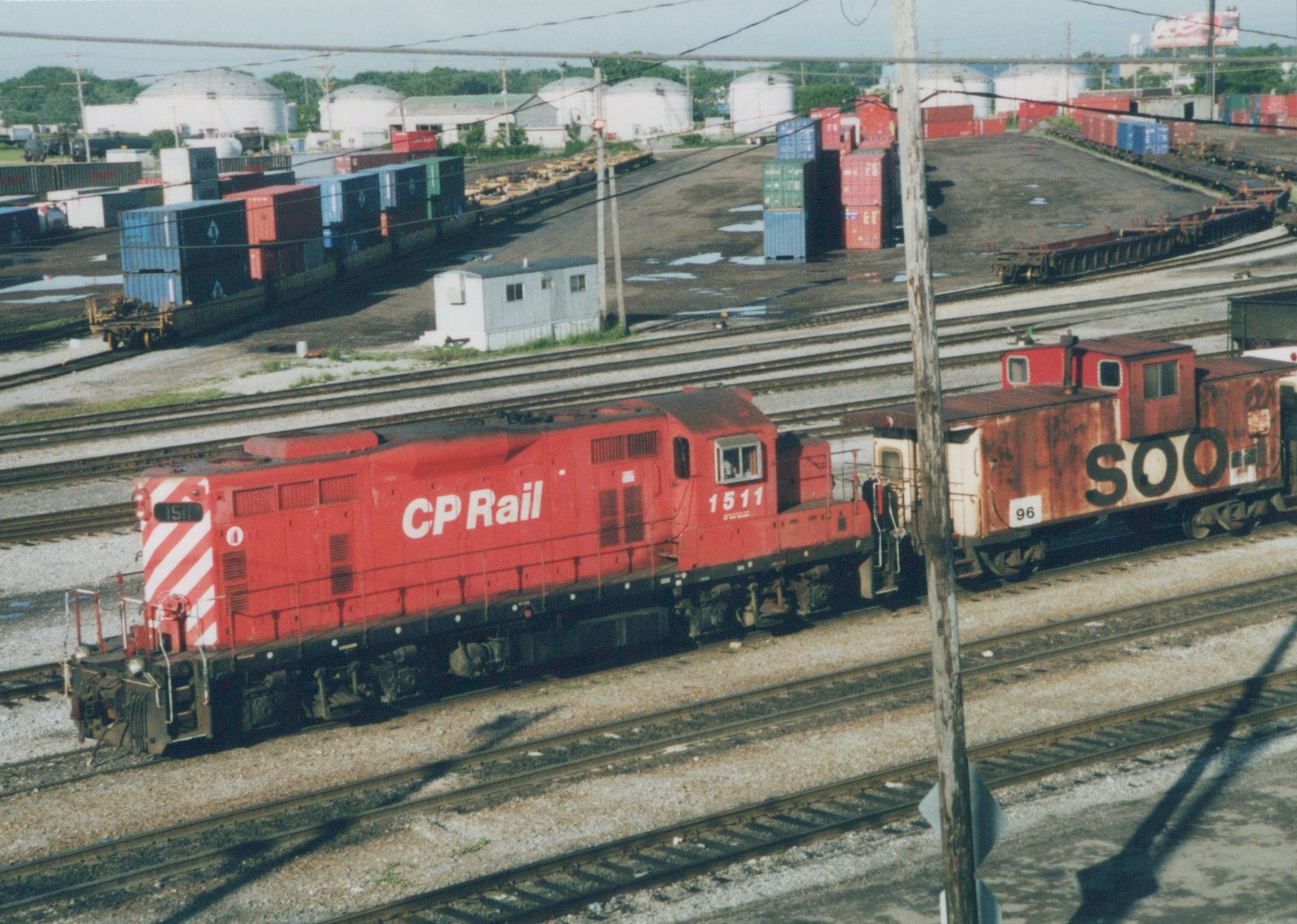 An overhead view of a red CP Rail locomotive numbered 1511 coupled to a weathered red Soo Line caboose numbered 96 on multiple tracks in a freight yard with shipping containers and tank farm in the background.